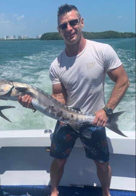 Photo de Stan Cappis qui tient un gros poisson et qui porte un short, un tee-shirt blanc et des lunettes de soleil. 