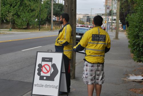 Two volunteers in yellow jackets stand outdoors beside a road with a sign showing a cartoon hand and a phone discouraging using a phone while driving