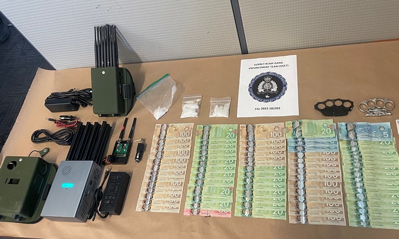 Photo Description: Four police radio jammers, an amount of cash featuring bills in values of $100, $50, $20, and $5, three small bags of what appears to be methamphetamine, and two sets of brass knuckles all on display on a table. 